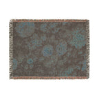 A rectangular woven throw blanket with fringed edges, featuring an abstract floral pattern in shades of brown, gray, and teal blue on a white background.