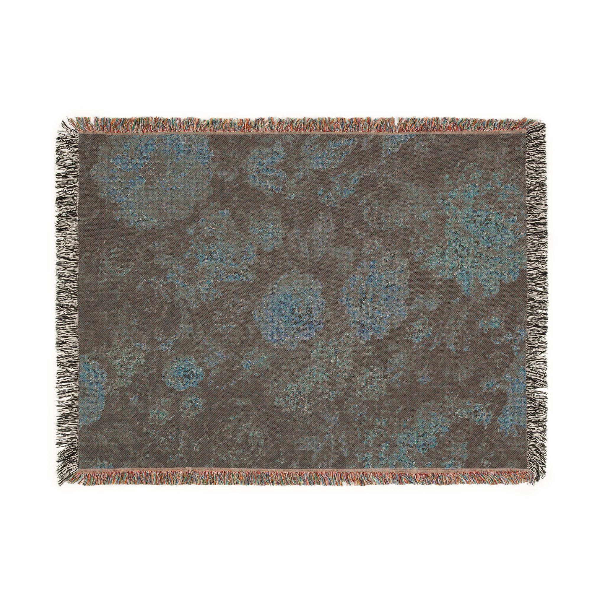 A rectangular woven throw blanket with fringed edges, featuring an abstract floral pattern in shades of brown, gray, and teal blue on a white background.