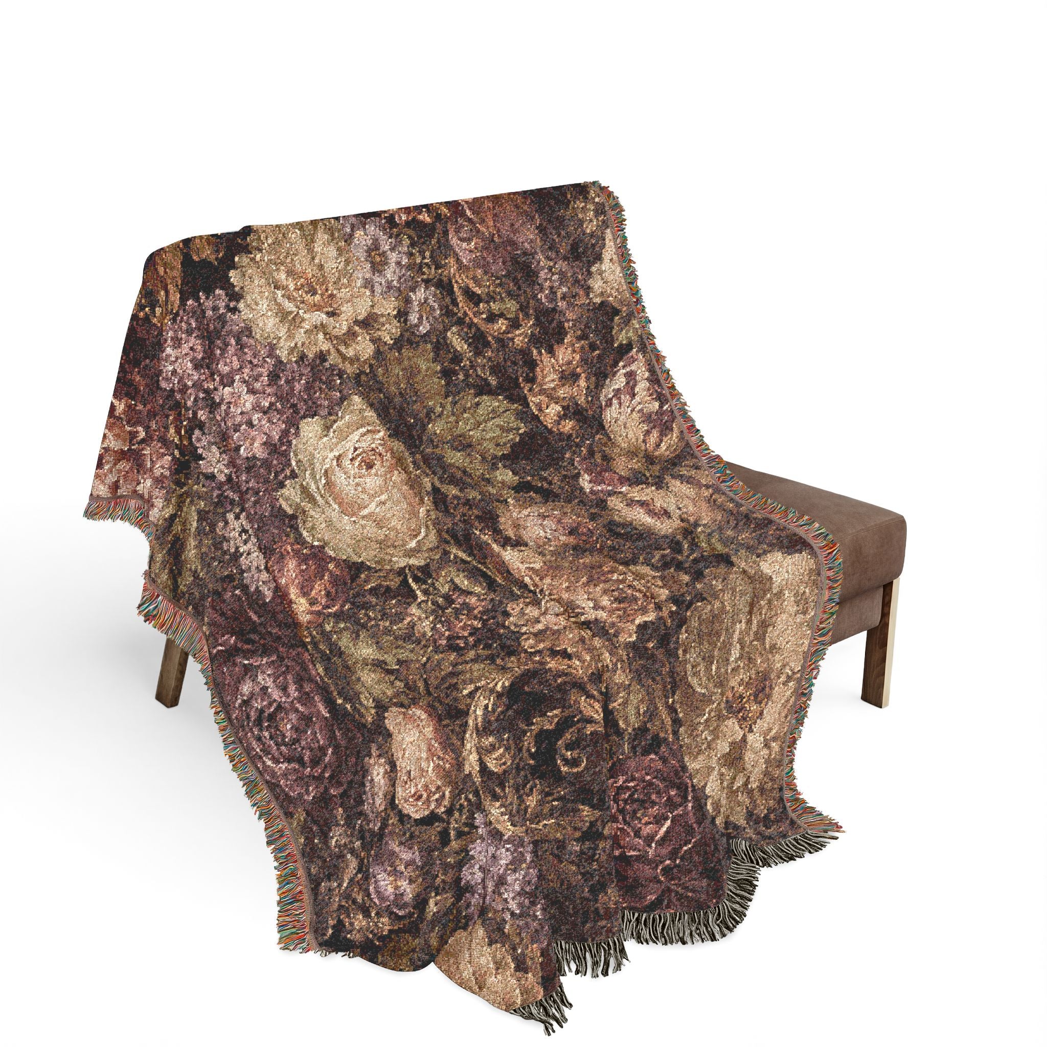 A fringed throw blanket with a vintage floral tapestry pattern in earthy brown, beige, and muted purple tones, draped over a brown upholstered bench against a white background.