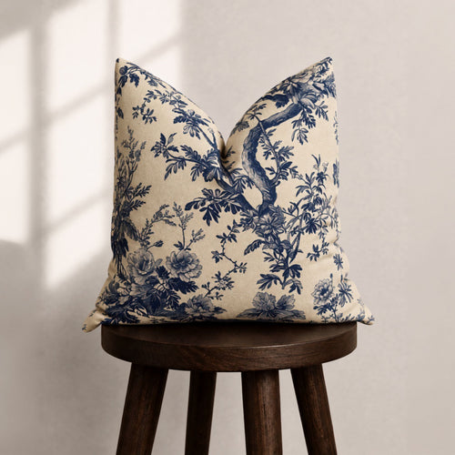 A square throw pillow with a navy blue floral and branch toile pattern on a cream background, displayed on a dark wooden stool against a neutral wall with soft window shadow light.