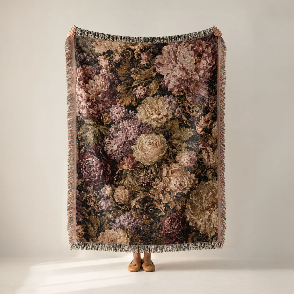 A person holds up a large woven throw blanket with a dense floral pattern in muted pink, beige, and brown tones, featuring fringed edges, against a plain light gray studio background.