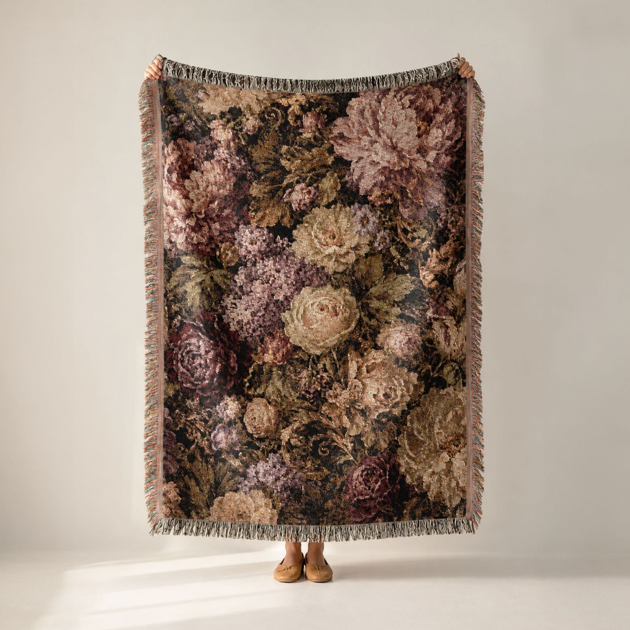 A person holds up a large woven throw blanket with a dense floral pattern in muted pink, beige, and brown tones, featuring fringed edges, against a plain light gray studio background.