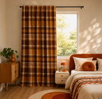 A tall window curtain in a warm brown and orange plaid pattern hanging on a black rod in a styled bedroom with a bed, nightstand, and wooden cabinet.