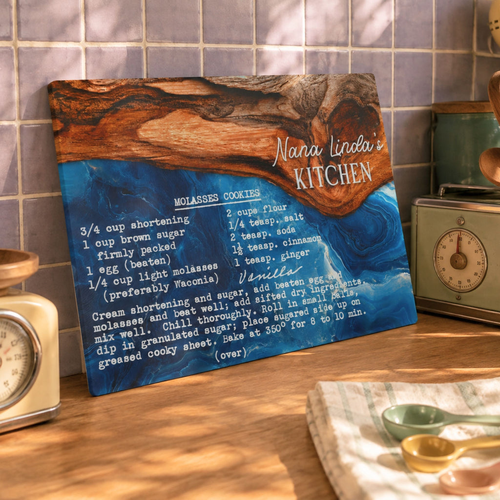 A personalized wooden recipe plaque leaning against a tiled kitchen wall, featuring a blue and brown resin design with the text 'Nana Linda's Kitchen' and a vanilla molasses cookie recipe printed in white. Surrounding kitchen props include a vintage-style clock, metal canister, and measuring spoons on a counter.