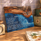 A personalized wooden recipe plaque leaning against a tiled kitchen wall, featuring a blue and brown resin design with the text 'Nana Linda's Kitchen' and a vanilla molasses cookie recipe printed in white. Surrounding kitchen props include a vintage-style clock, metal canister, and measuring spoons on a counter.
