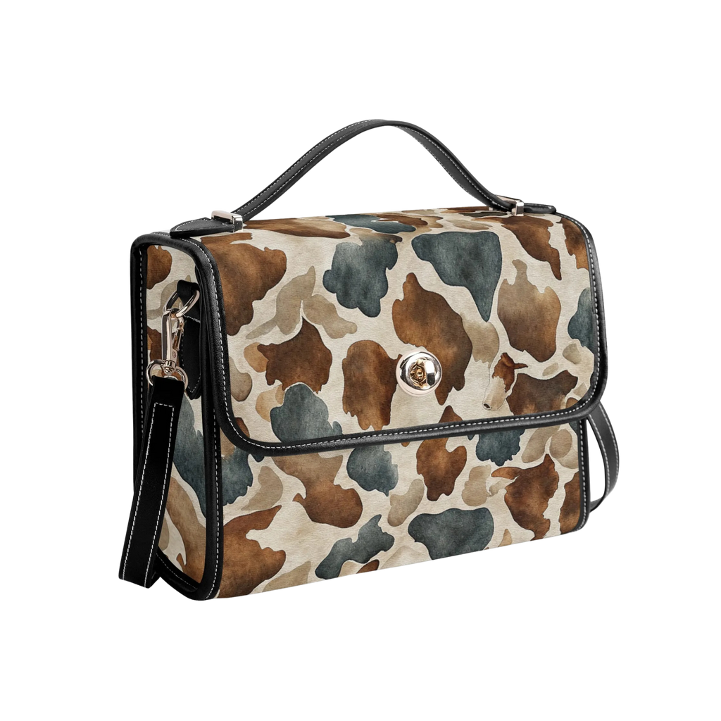A structured handbag with a brown, beige, and gray watercolor cow print pattern, featuring black leather trim, a top handle, and a front flap with a silver-tone snap button.