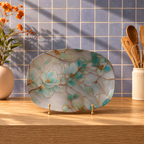 An oval ceramic serving platter with a white marble-look base and a blue floral pattern outlined in gold, displayed on a wooden table in a kitchen setting with a blue tiled backsplash.