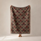 A person holds up a large woven throw blanket with a vintage-style floral damask pattern in muted red, beige, and black, with fringed edges, against a neutral beige background.