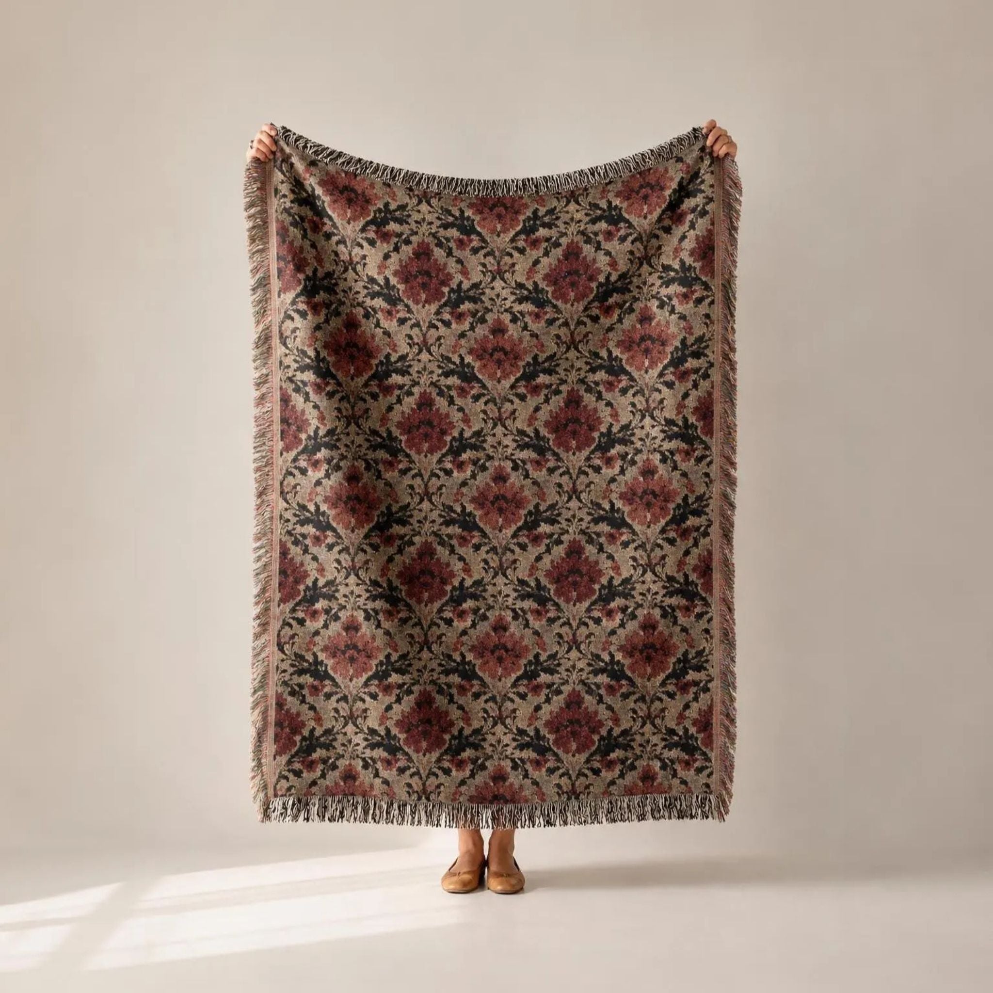 A person holds up a large woven throw blanket with a vintage-style floral damask pattern in muted red, beige, and black, with fringed edges, against a neutral beige background.