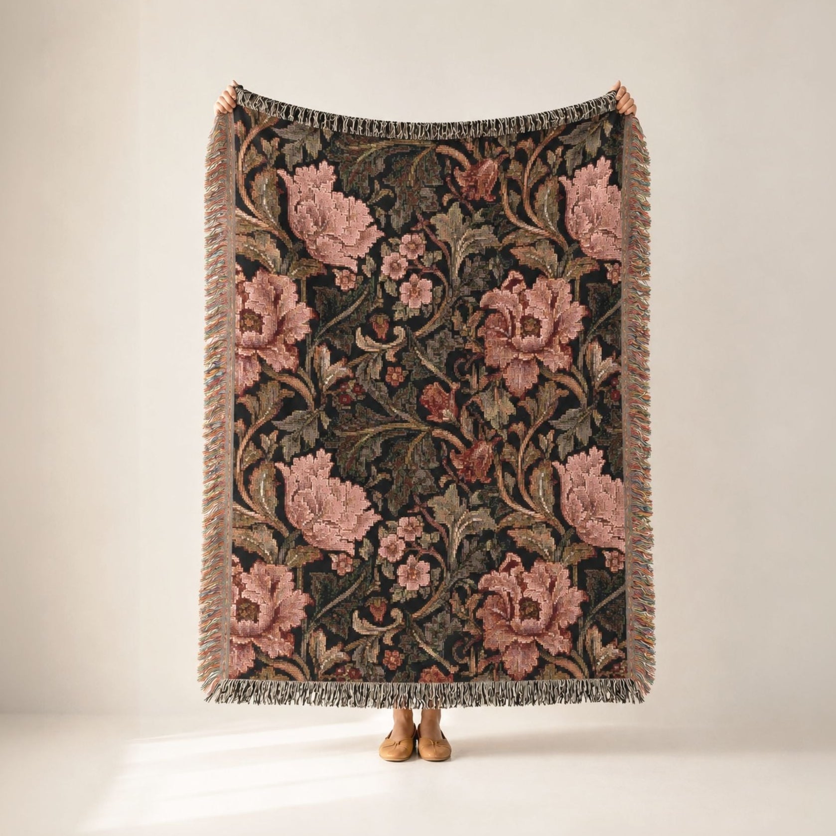 A rectangular woven throw blanket with fringe, featuring a dark floral tapestry pattern with large pink flowers and green leaves, held up by a person whose feet are visible at the bottom against a plain light beige studio background.