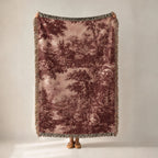A person holds up a large woven throw blanket with a muted forest pattern in dusty rose and brown tones, featuring fringed edges, against a plain light gray studio background.