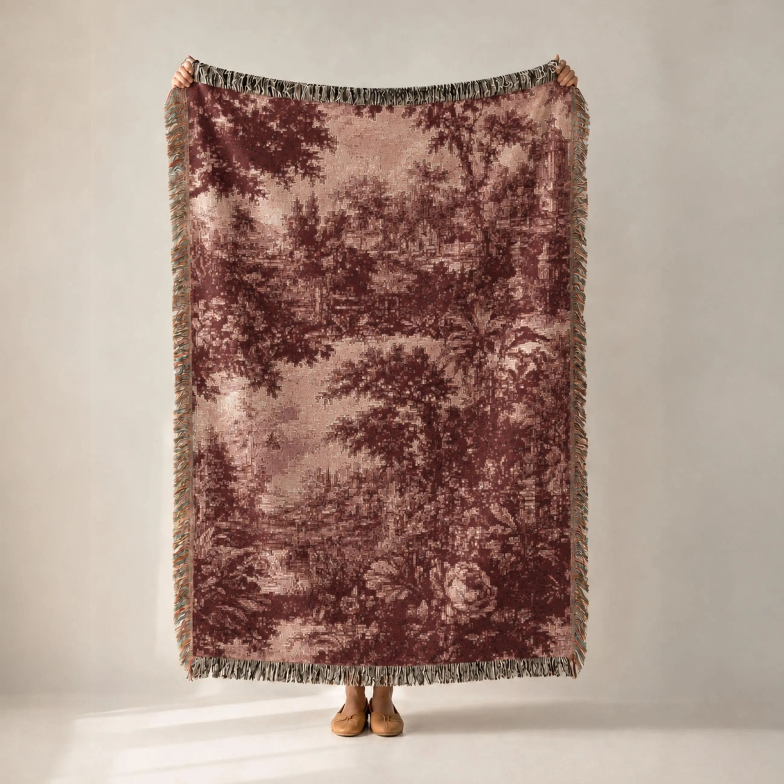 A person holds up a large woven throw blanket with a muted forest pattern in dusty rose and brown tones, featuring fringed edges, against a plain light gray studio background.