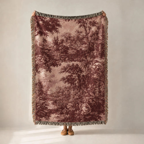 A person holds up a large woven throw blanket with a muted forest pattern in dusty rose and brown tones, featuring fringed edges, against a plain light gray studio background.