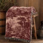 A plush velvet throw blanket with a faded landscape print in burgundy and dusty pink, draped over a rustic wooden stool in a cozy room setting.