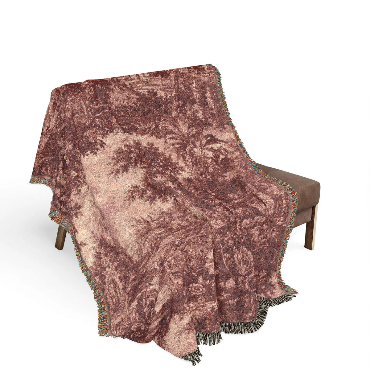 A woven throw blanket with a classic toile landscape print in dusty rose and brown, draped over a brown armchair against a solid white background.