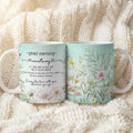 Two ceramic mugs with floral designs and personalized text, displayed side-by-side on a cozy cream-colored knit blanket. One mug shows a white background with black cursive text reading 'good morning - courtney' and floral accents, while the other shows a mint green background with detailed botanical illustrations.