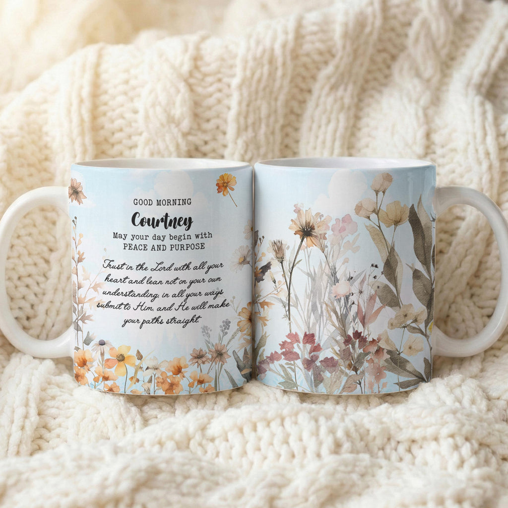 Two ceramic mugs with floral illustrations and a morning greeting printed on them, displayed side by side on a soft cream knit blanket.