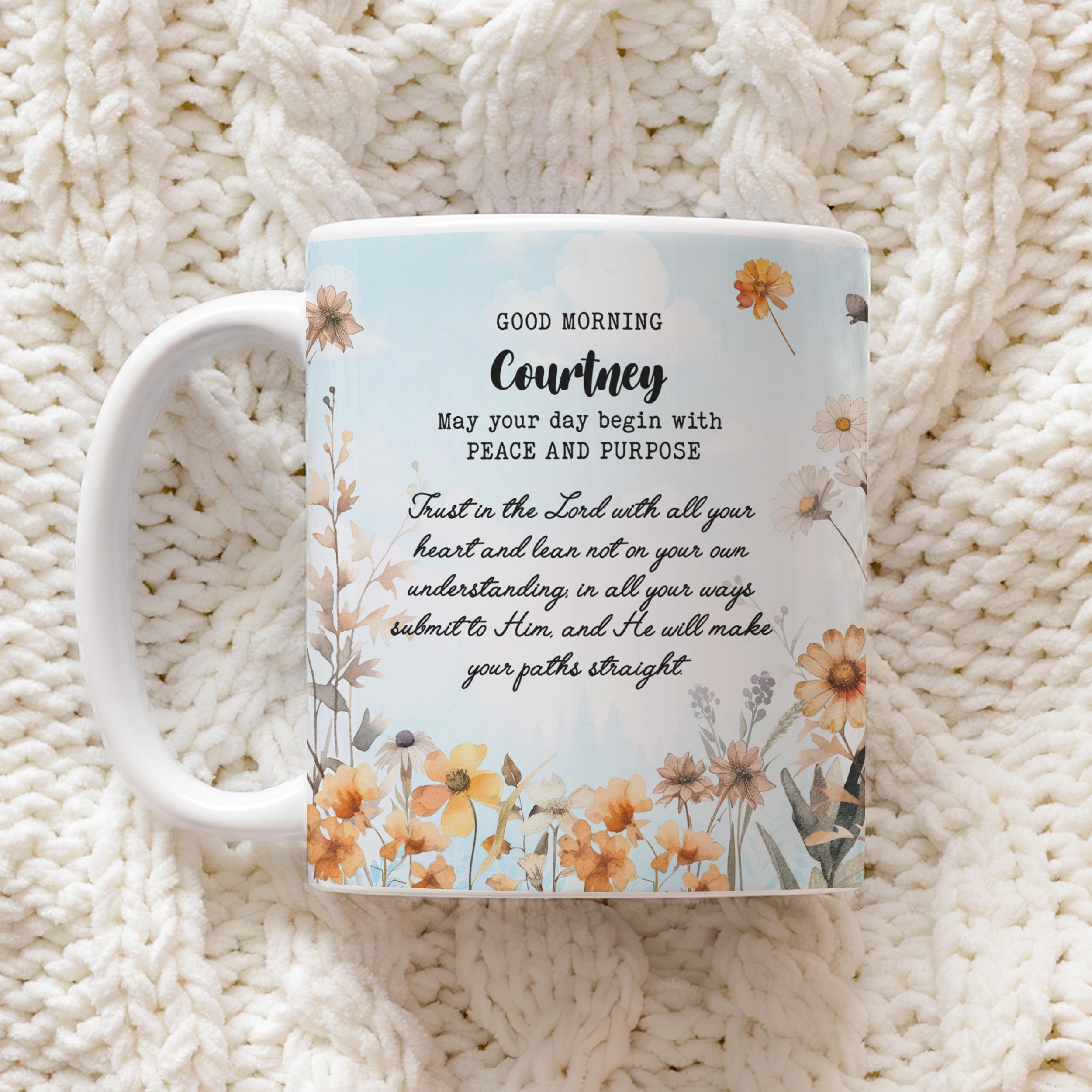 A white ceramic mug with a floral design and a morning prayer printed on it, resting on a textured cream-colored knit blanket.