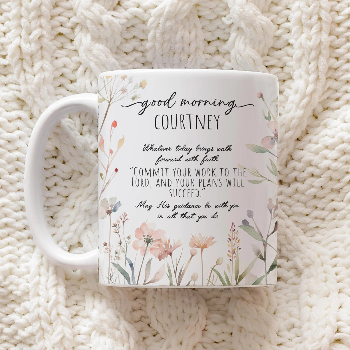 A white ceramic mug with a personalized Bible verse and floral illustration, displayed on a cozy cream-colored chunky knit blanket.
