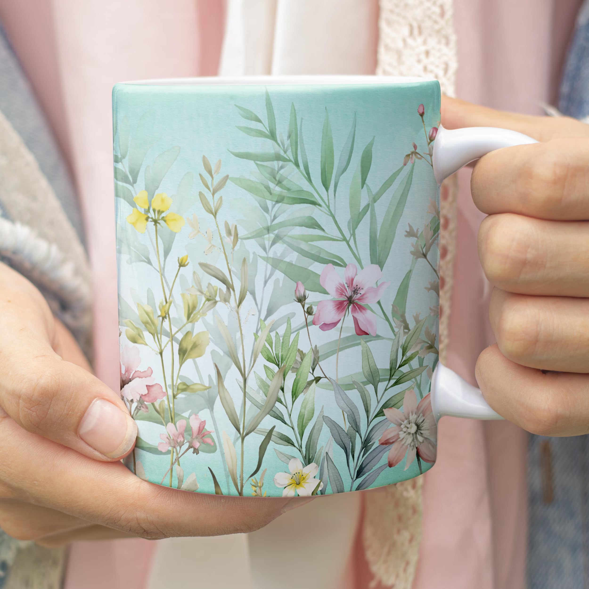 A person holding a square ceramic mug with a watercolor-style botanical print featuring green leaves and pink, yellow, and white flowers on a pale aqua background.