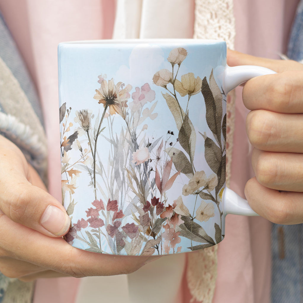 A white ceramic mug with a watercolor floral design, held by two hands against a soft pink and beige background.