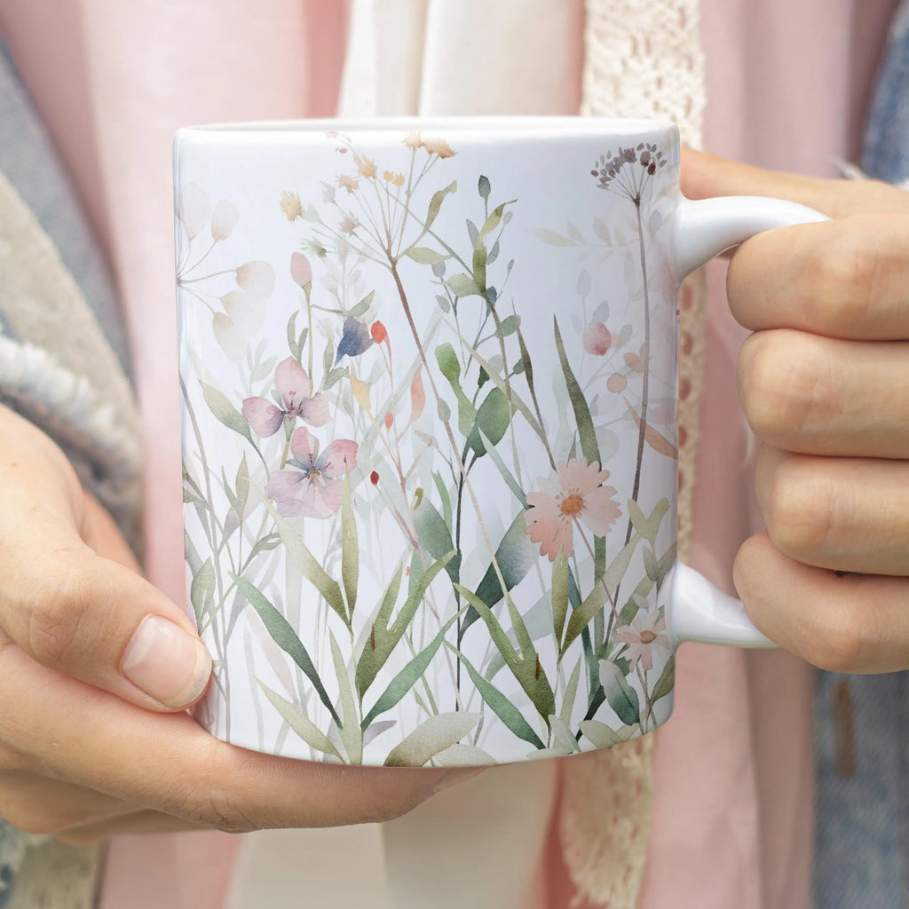 A white ceramic mug with a delicate watercolor wildflower and botanical pattern, held by two hands.