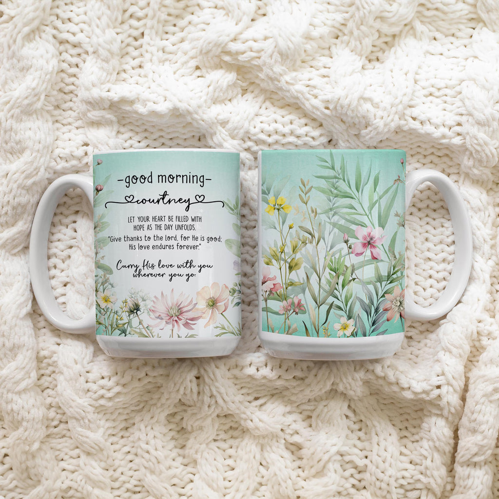 Two white ceramic mugs with floral and text designs, displayed side by side on a cream-colored cable-knit blanket. The left mug features a 'Good Morning Courtney' quote with pink flowers, and the right mug features a light green botanical pattern with pink and yellow flowers.