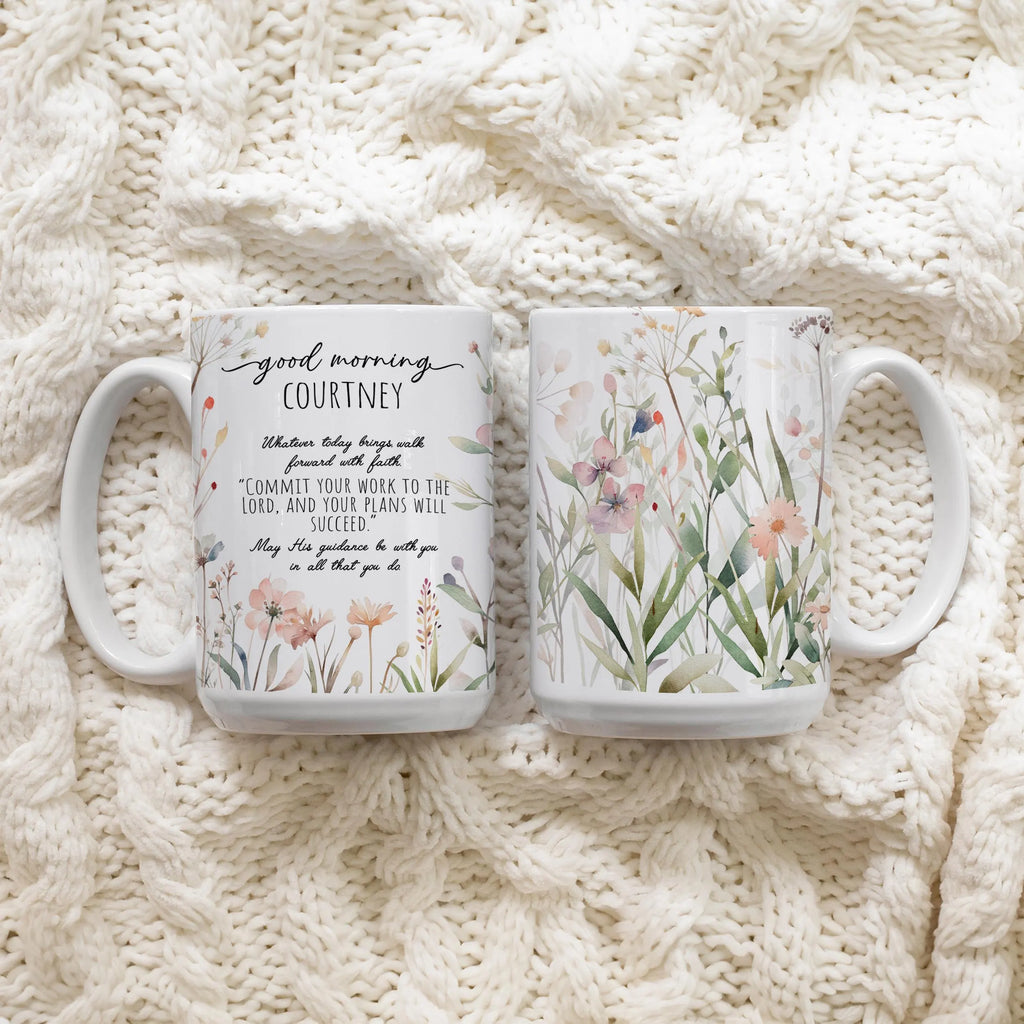 Two white ceramic mugs with floral and text designs featuring the front and back of the mug, placed on a cream cable-knit blanket.
