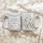 Two white ceramic mugs with floral and text designs featuring the front and back of the mug, placed on a cream cable-knit blanket.