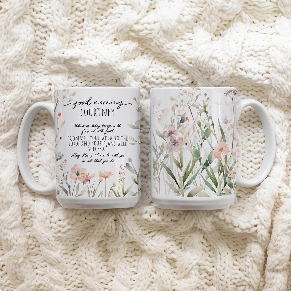 Two white ceramic mugs with floral and text designs featuring the front and back of the mug, placed on a cream cable-knit blanket.