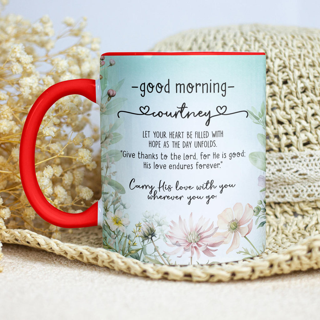 A personalized ceramic mug with a red handle and rim, featuring a light blue floral design and a morning greeting quote for 'courtney'. The mug is styled on a beige woven mat with dried flowers in the background.