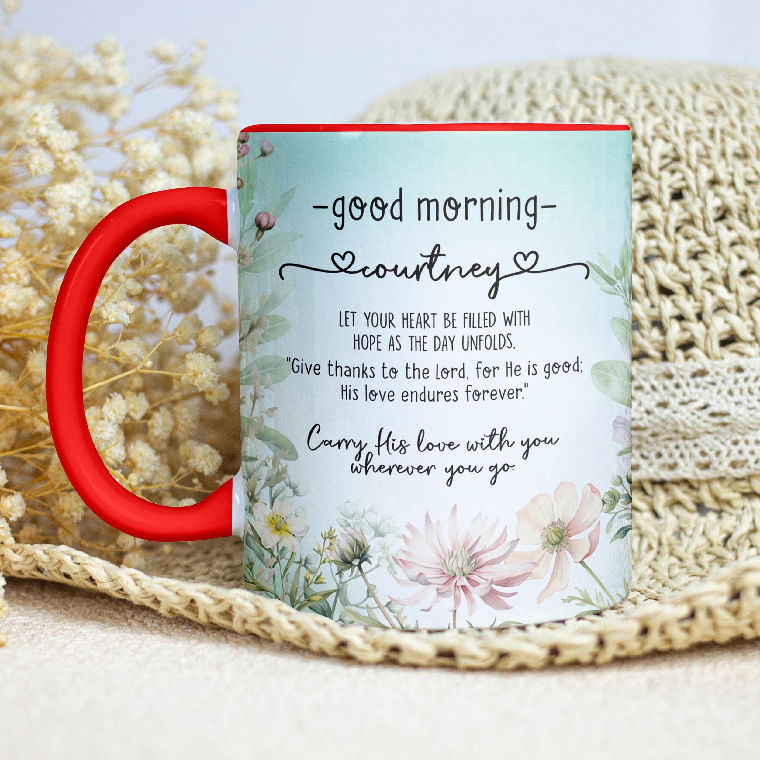 A personalized ceramic mug with a red handle and rim, featuring a light blue floral design and a morning greeting quote for 'courtney'. The mug is styled on a beige woven mat with dried flowers in the background.