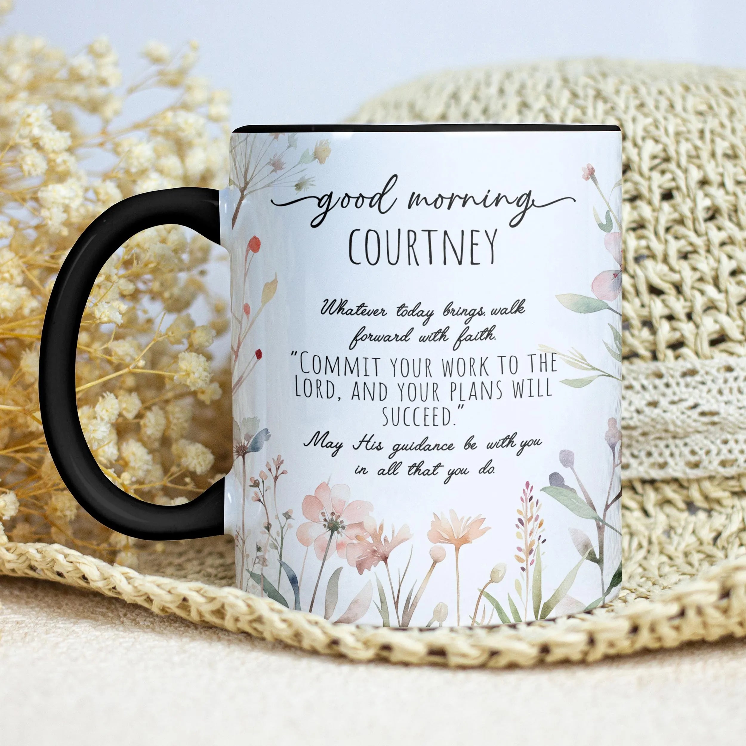 A ceramic mug with a black handle and black rim, printed with a floral design and a morning prayer message for 'Courtney'. The mug is styled on a beige woven mat with dried flowers in the background.