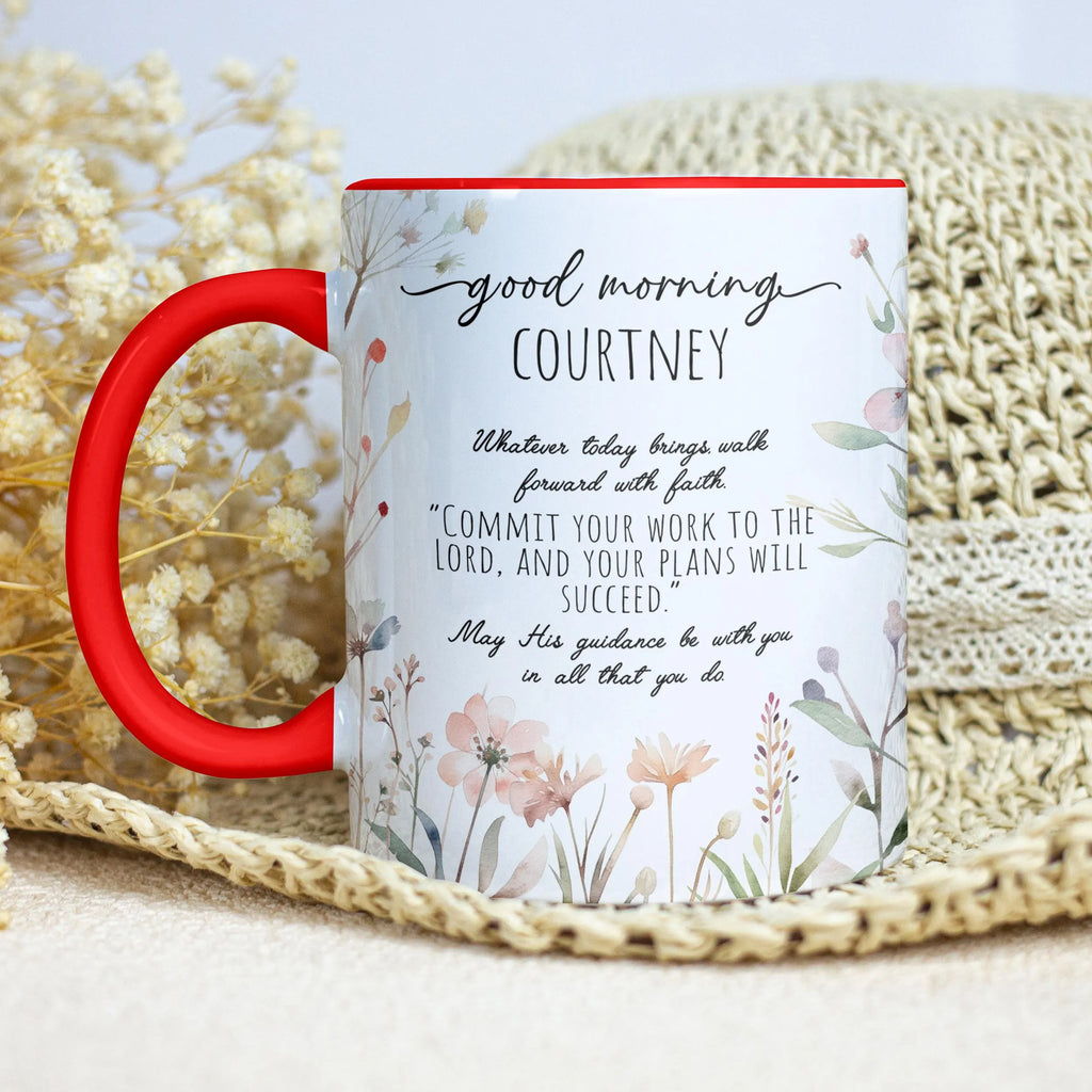 A personalized ceramic mug with a red handle and red rim, featuring a floral design and a Bible verse printed on the side. The mug is styled on a beige woven mat with dried flowers in the background.