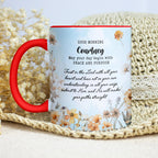 A personalized ceramic mug with a red handle and red rim, featuring a light blue printed design with floral illustrations and a morning prayer for 'Courtney'. The mug is styled on a beige woven mat with dried flowers in the background.