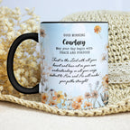 A ceramic mug with a black handle and black rim, featuring a light blue floral design with a Bible verse and the name 'Courney' printed on it. The mug is styled on a beige woven mat with dried white flowers in the background.